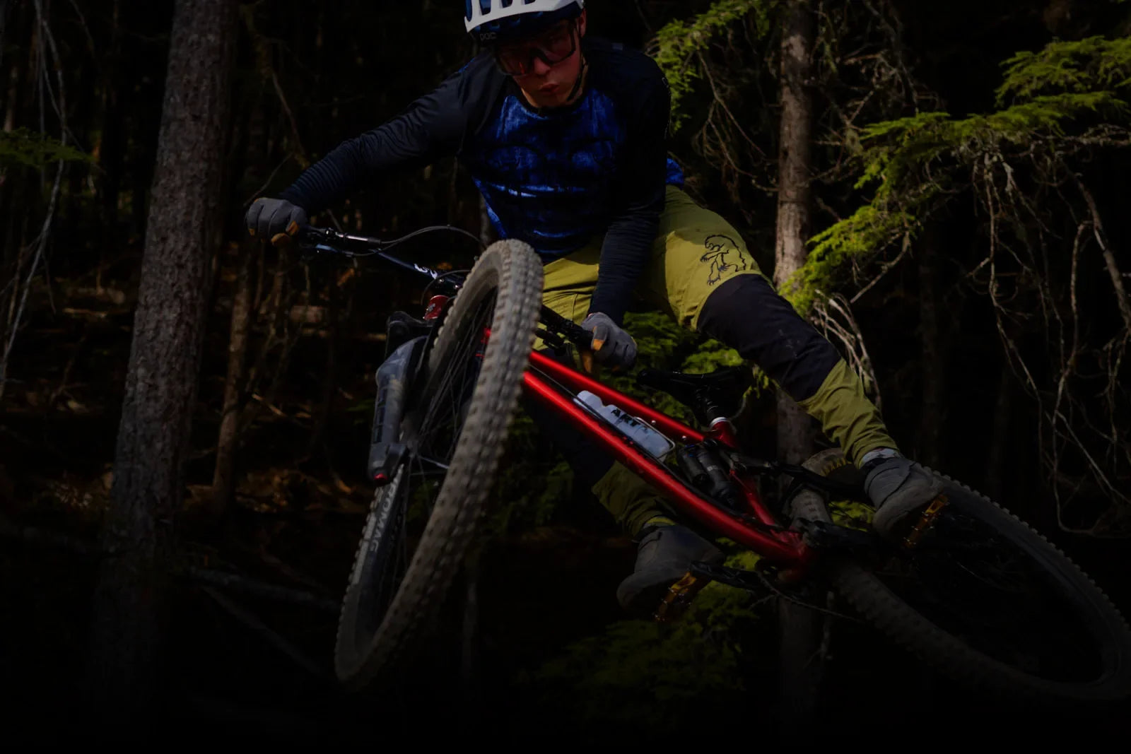 A mountain biker in a helmet and protective gear is captured mid-air on a red mountain bike, against a dark, dense forest background. The biker is looking towards the camera with a focused expression.