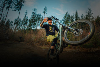 A man on a mountain bike performs a wheelie on a dirt path, smiling and waving at the camera. Tall pine trees and bare deciduous trees line the background under a bright sky.