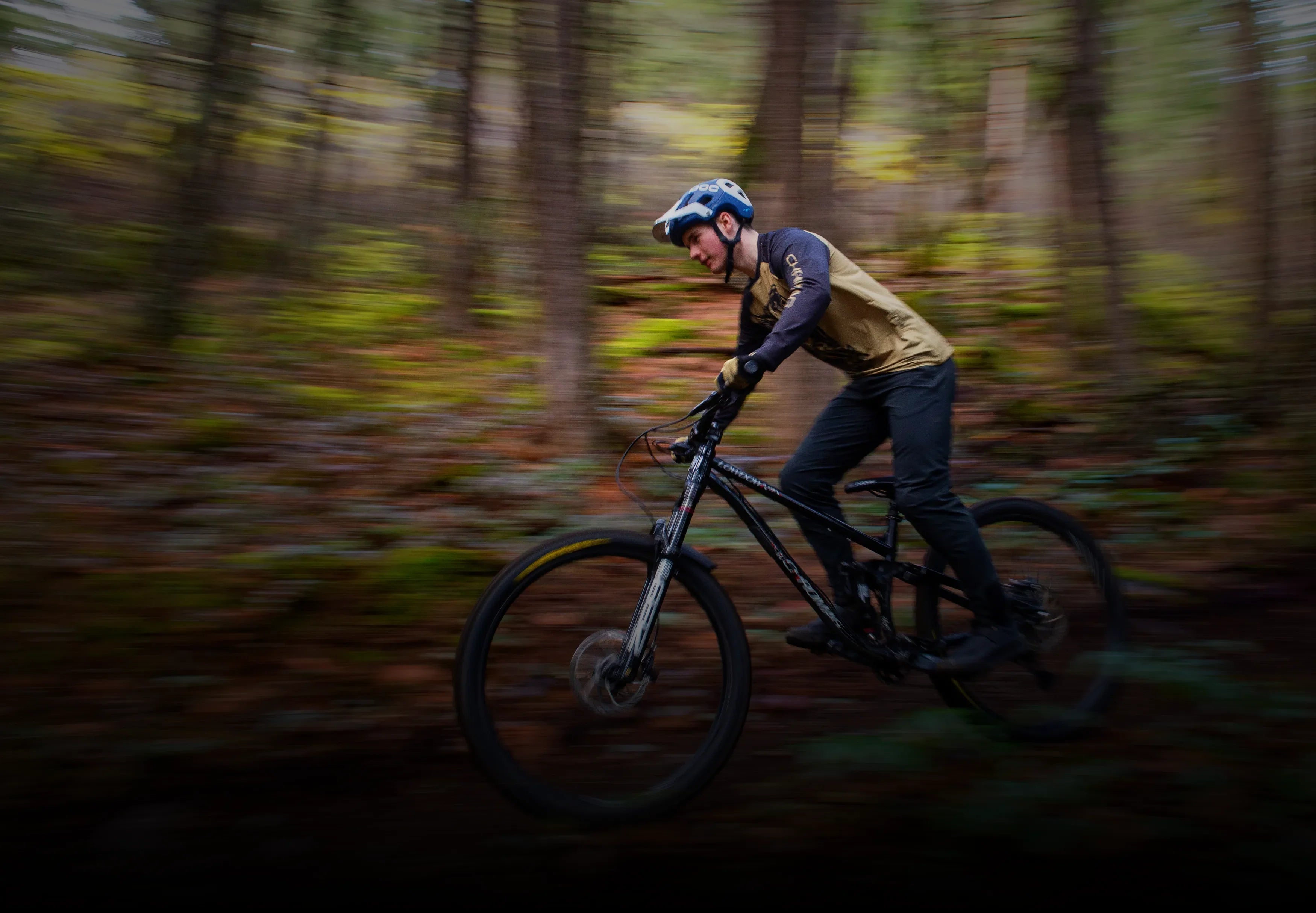 A mountain biker in a helmet and long-sleeved jersey rides a black full-suspension mountain bike through a forest trail. The background is blurred to convey motion, with tall trees and fallen leaves visible.