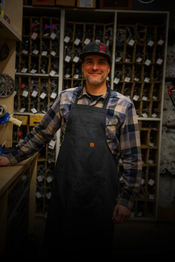 A man in a plaid shirt, black apron, and baseball cap smiles at the camera, leaning on a wooden workbench. Behind him are shelves filled with various parts and components.