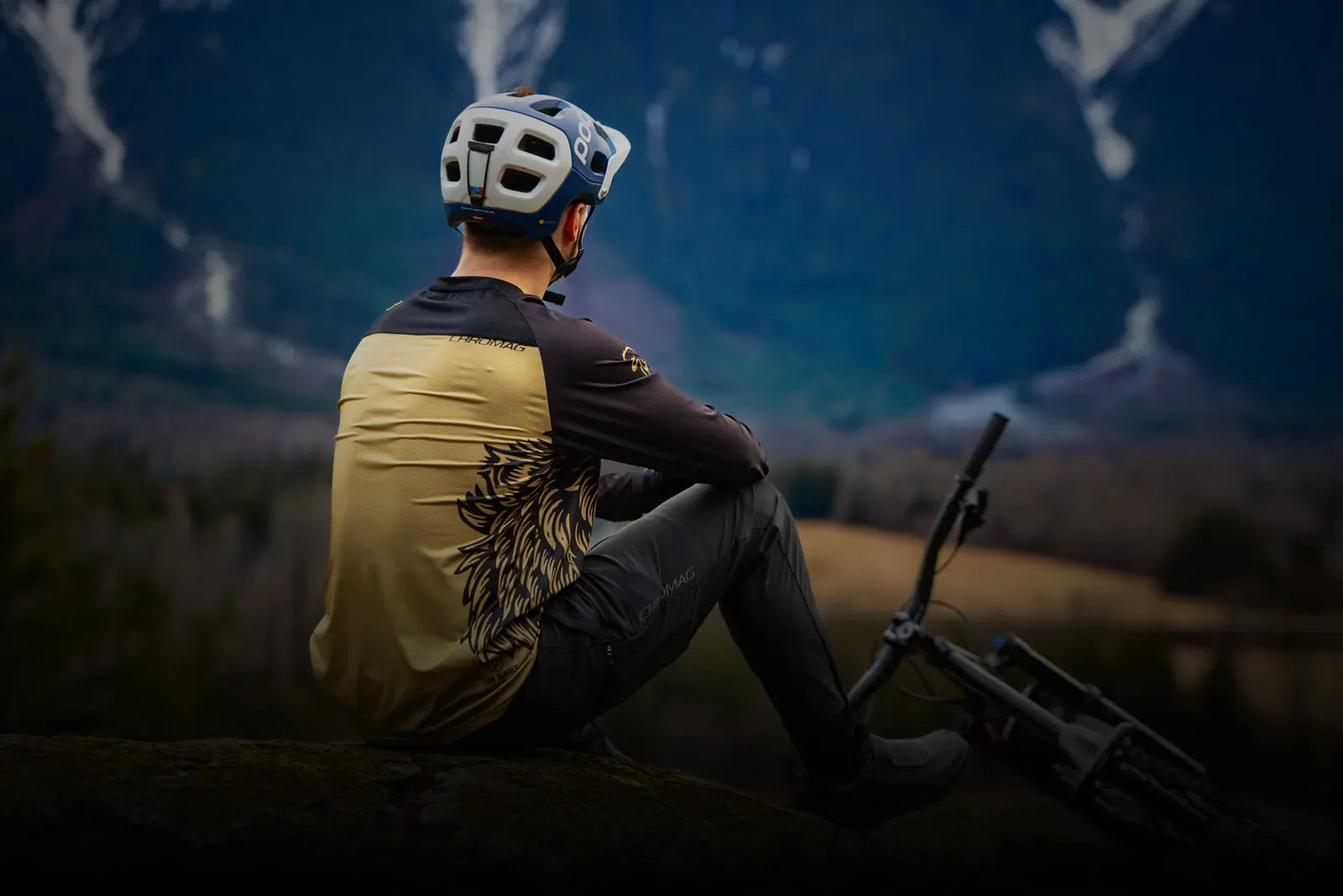 A person wearing a long-sleeved cycling jersey with a graphic design and dark pants, sitting on a rock next to a mountain bike, looking out at a blurred mountain and forest landscape.