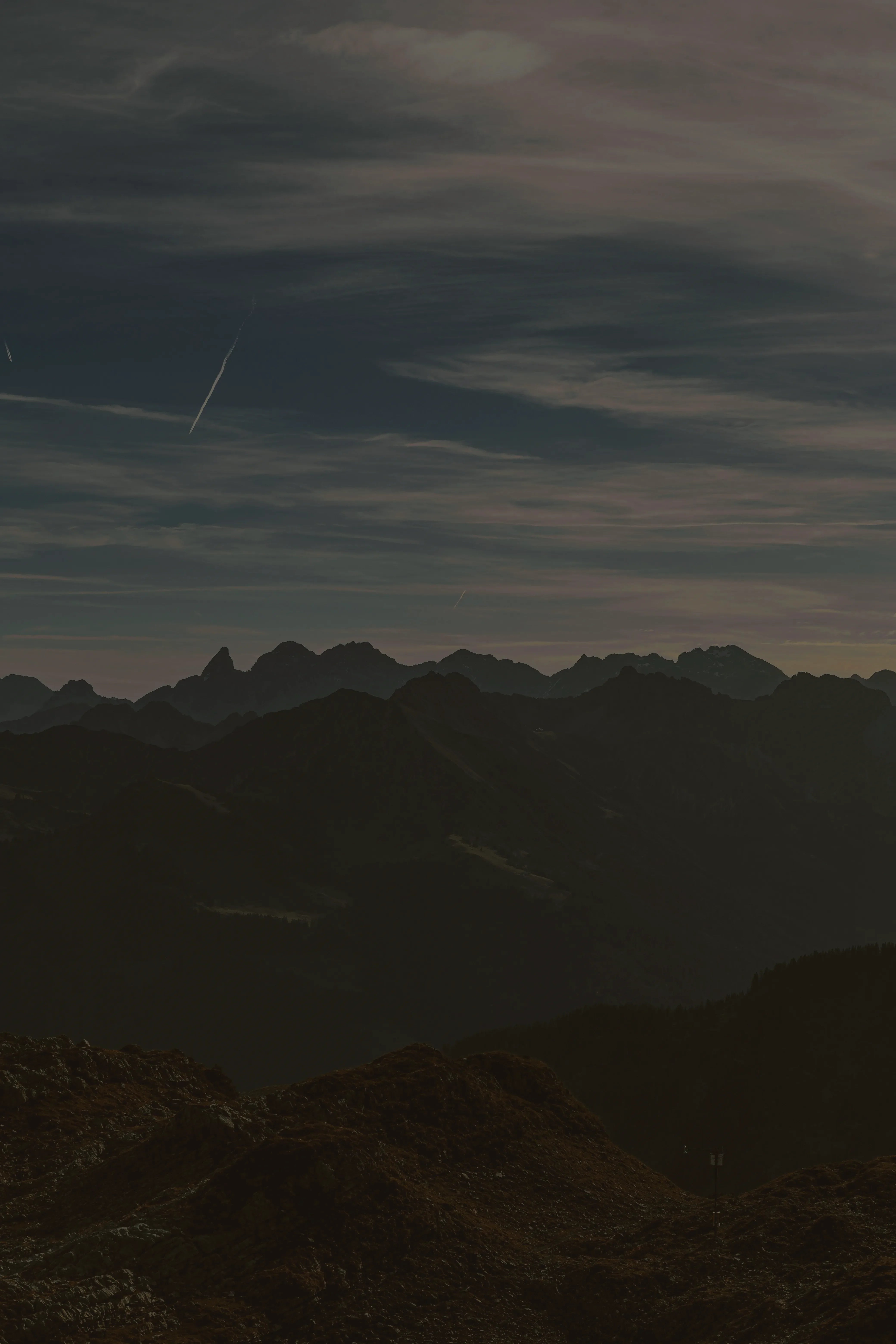 A vast mountain range with dark peaks and valleys stretches under a partly cloudy sky, with faint contrails visible. The foreground features a rocky, grass-covered hill in muted earth tones.